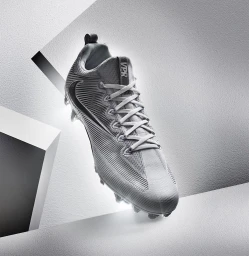 A grey Nike sports cleat positioned diagonally against a textured white architectural background with dramatic studio lighting.