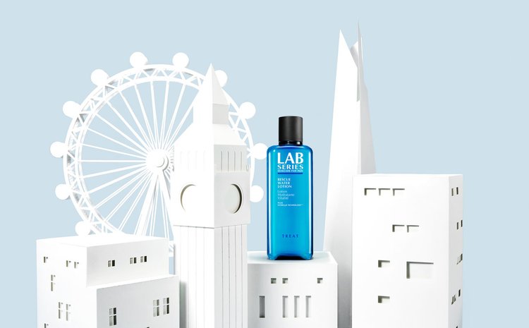 Lab Series skincare product bottle stands prominently in front of white paper models of London landmarks, set against a light blue background.