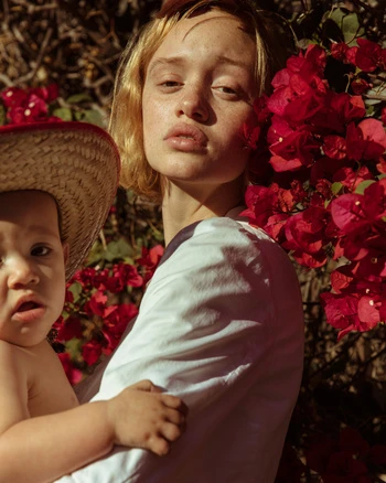 Portrait of a young woman holding a toddler in a straw hat, with vibrant red bougainvillea flowers in the background, in natural light.