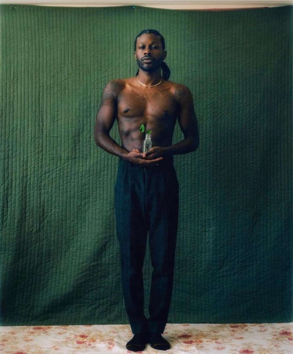 Shirtless Black man with braided hair holding a small glass bottle against a dark green quilted backdrop by Bryan Rodner Carr
