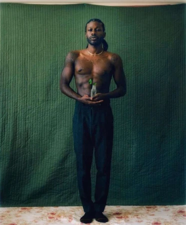 Shirtless Black man with braided hair holding a small glass bottle against a dark green quilted backdrop by Bryan Rodner Carr