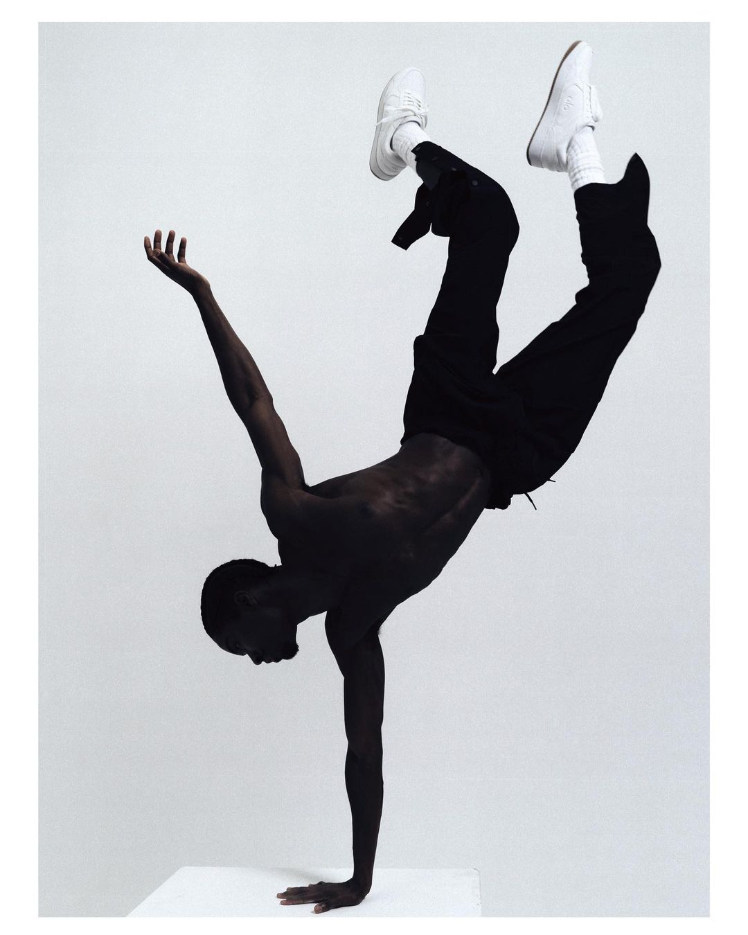 African American man in a handstand wearing sneakers, black pants, against a light grey background in a studio setting.