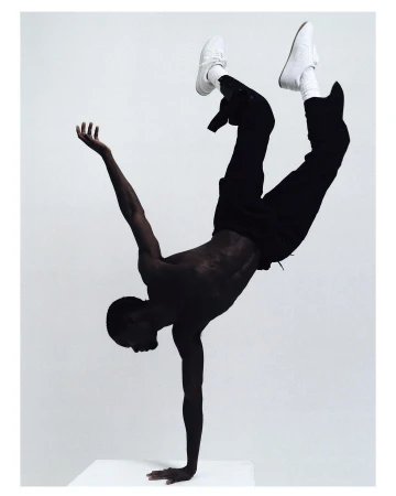African American man in a handstand wearing sneakers, black pants, against a light grey background in a studio setting.