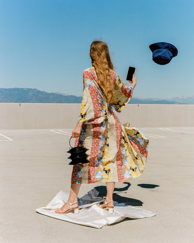 Back view of a woman in a floral robe on a rooftop, holding a phone with a hat floating in the air, against a mountain backdrop.