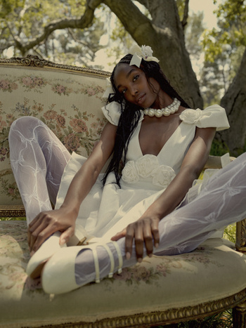 Elegant woman in a white dress and tights poses on an ornate sofa in a natural outdoor setting with trees in the background.