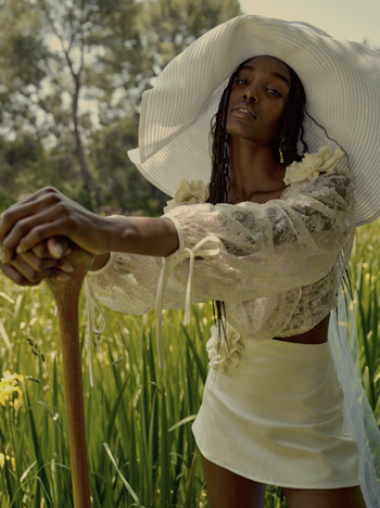 Elegant Black woman poses in a sunlit field of tall grasses, wearing a large white hat, sheer blouse, and cream skirt, holding a gardening tool.