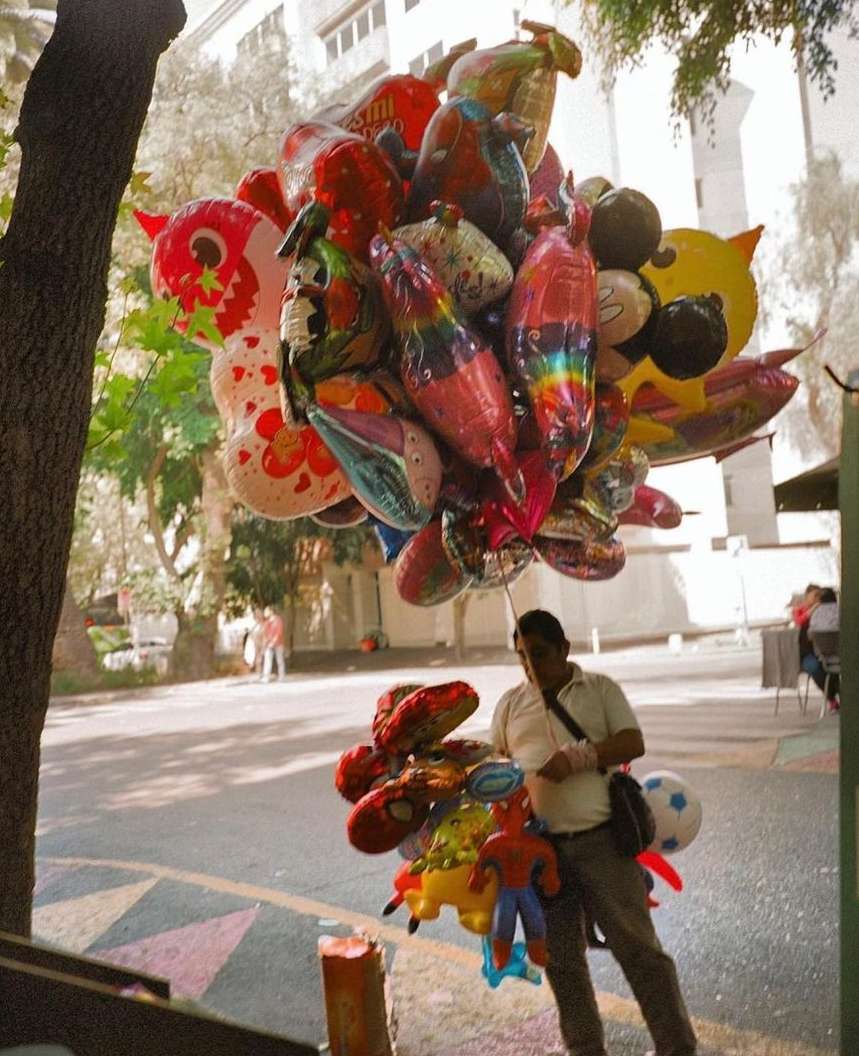 Outdoor shot of a man selling character balloons in an urban setting, brightly colored balloons fill the frame with pedestrian sidewalk.