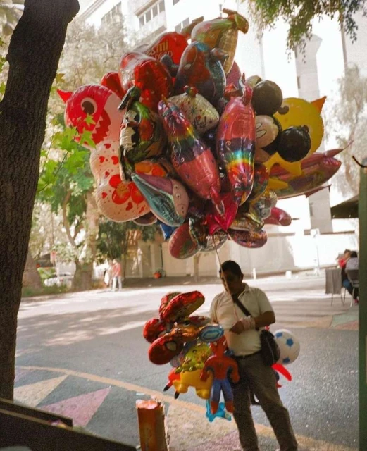 Outdoor shot of a man selling character balloons in an urban setting, brightly colored balloons fill the frame with pedestrian sidewalk.