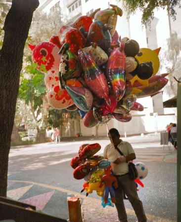 Outdoor shot of a man selling character balloons in an urban setting, brightly colored balloons fill the frame with pedestrian sidewalk.