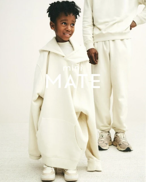Child model poses in oversized cream-colored hooded sweatshirt and matching sneakers next to an adult in a matching outfit.