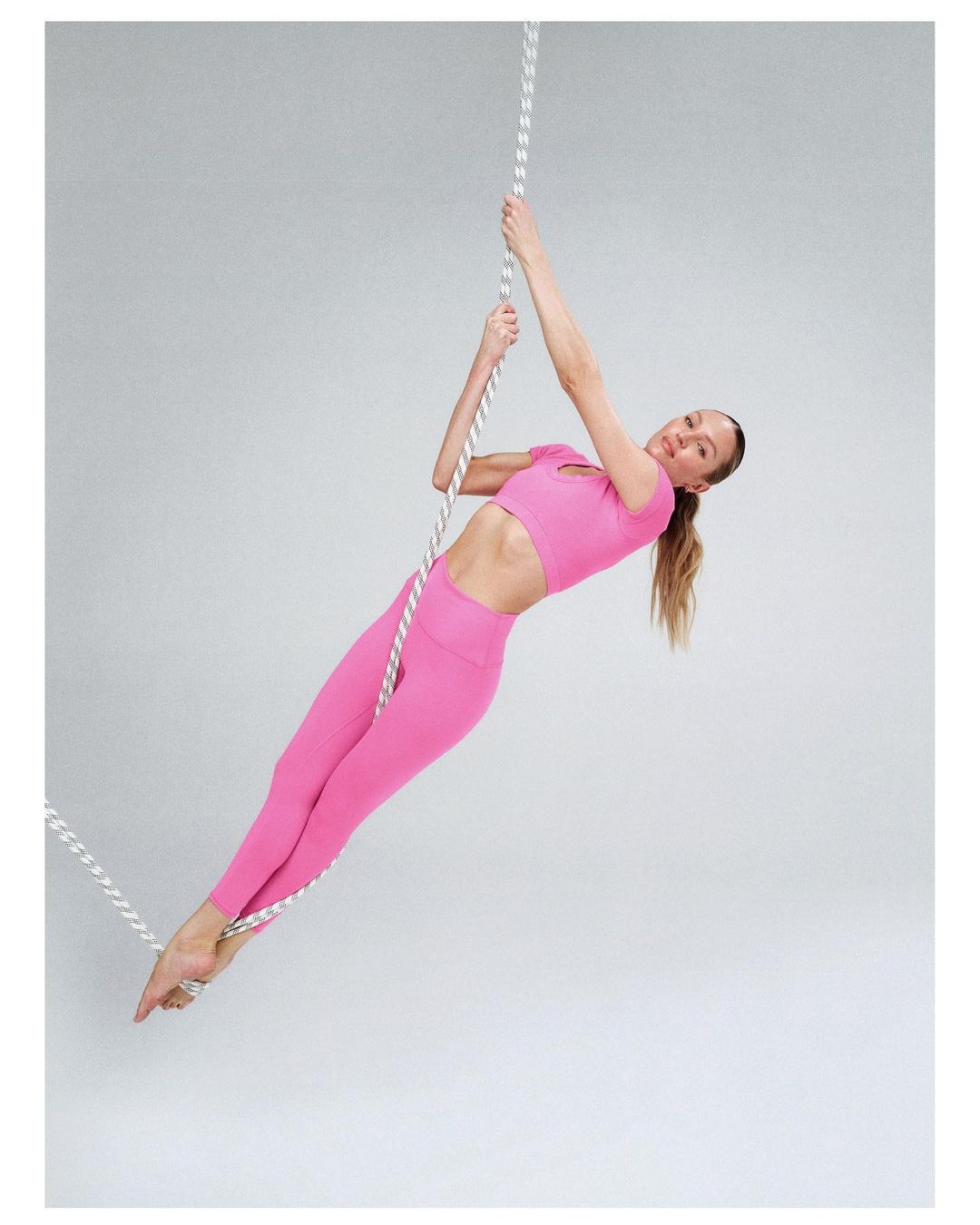 A woman in pink athletic wear poses while hanging from a climbing rope in a brightly lit photo studio against a gray background.