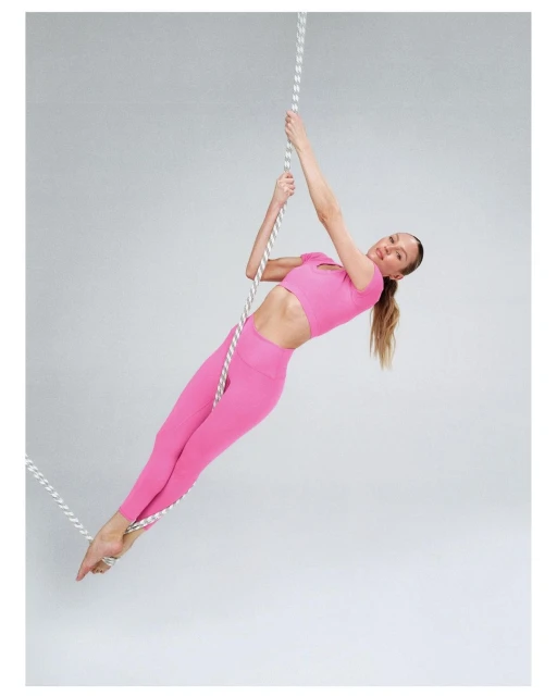 A woman in pink athletic wear poses while hanging from a climbing rope in a brightly lit photo studio against a gray background.