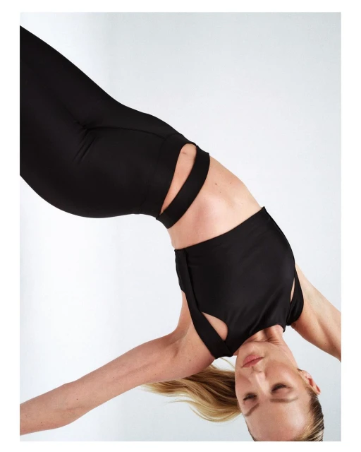 A woman in black workout gear is bending over backward against a plain white backdrop in a studio setting, showcasing activewear fashion.