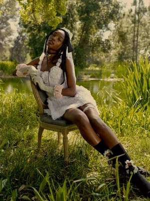 Woman in white floral dress poses on chair in garden. Outdoor setting with lush greenery and pond backdrop. Floral accents and elegant chair.
