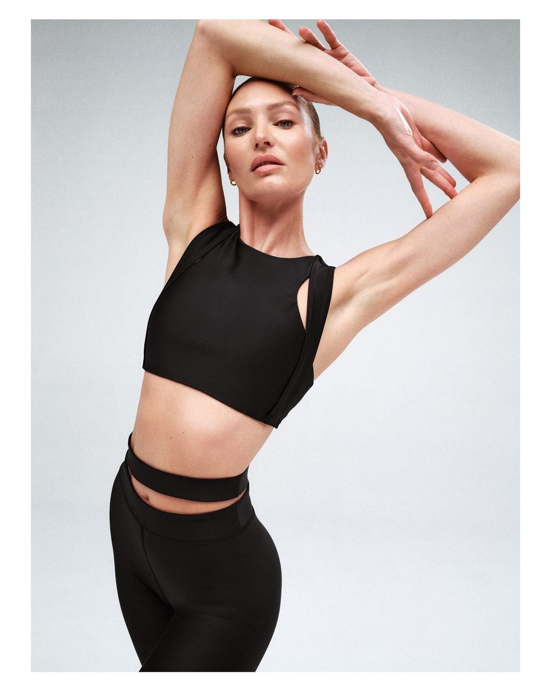 A slender, tan-skinned woman in black athletic top and leggings poses with her arms raised in a studio setting, showcasing fitness apparel.