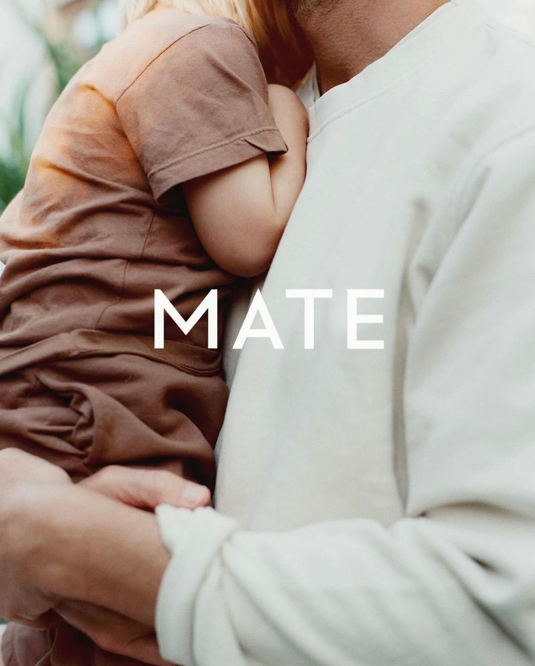 Adult holding a small child wearing brown clothing with the word MATE overlaid