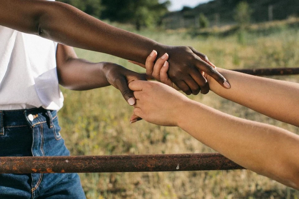 Close-up of two light-skinned and one dark-skinned person holding hands over a rusty metal bar in a field of dry grass.