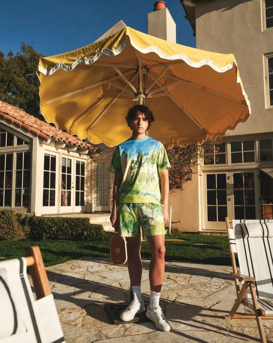 A young adult poses outdoors in patterned shorts and tee, holding a paddle, under a yellow umbrella, with a building backdrop.