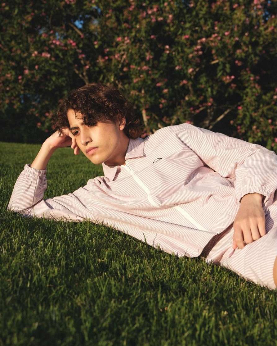 Young man in striped casual outfit reclines on grass outdoors with flowering tree in background on a sunny day, eye-level perspective.