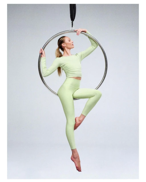 Athletic woman in a light green bodysuit performing aerial hoop on a silver ring, suspended in a bright grey, evenly lit space.