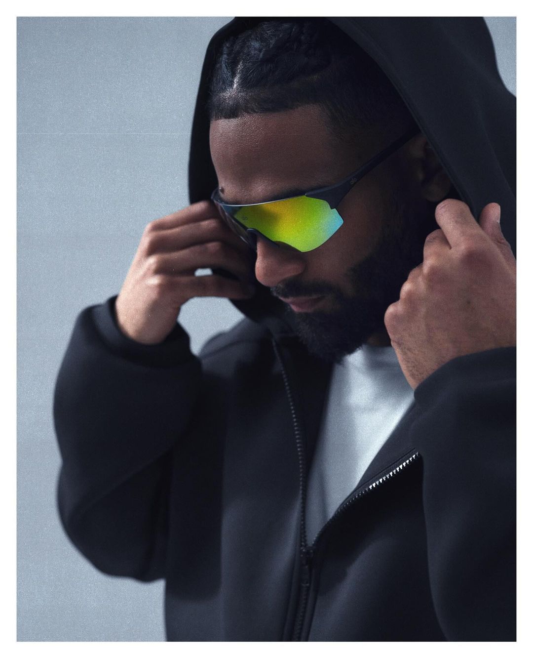An adult male wearing mirrored sport sunglasses and a black hooded jacket, looking down with hands adjusting the hood in a studio shot.
