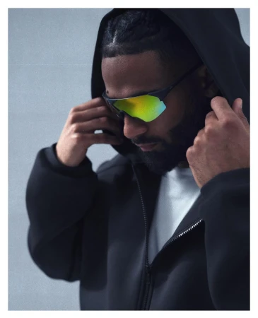 An adult male wearing mirrored sport sunglasses and a black hooded jacket, looking down with hands adjusting the hood in a studio shot.