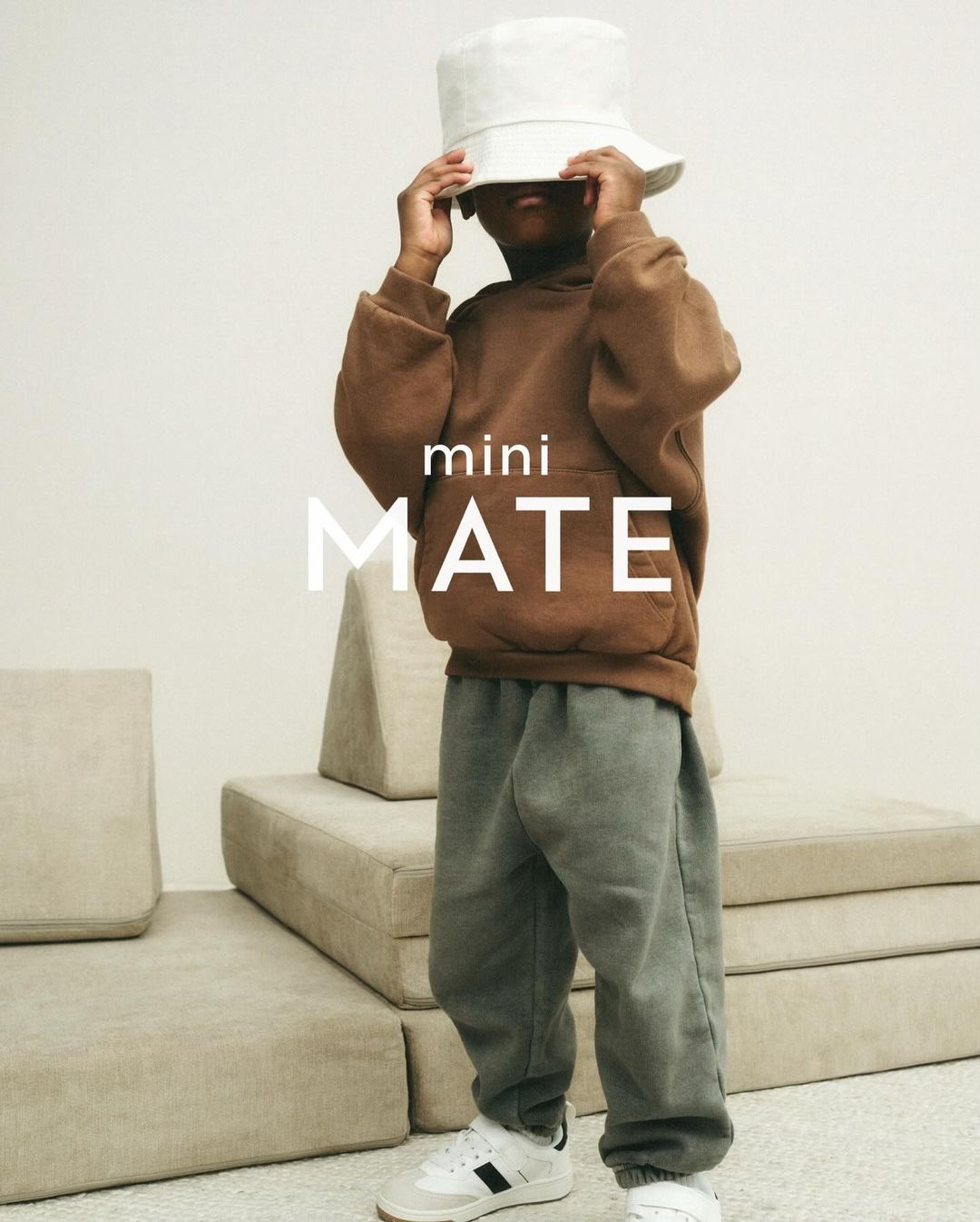 Child in brown hoodie and grey sweatpants stands in front of tan backdrop wearing a white bucket hat indoors.