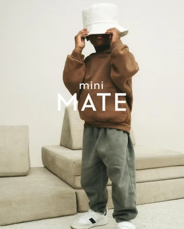 Child in brown hoodie and grey sweatpants stands in front of tan backdrop wearing a white bucket hat indoors.