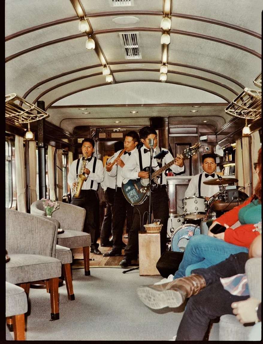 Live band performs in a train car with tables and chairs; musicians playing saxophone, flute, guitar, and drums, and listeners sit nearby.