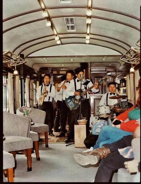 Live band performs in a train car with tables and chairs; musicians playing saxophone, flute, guitar, and drums, and listeners sit nearby.