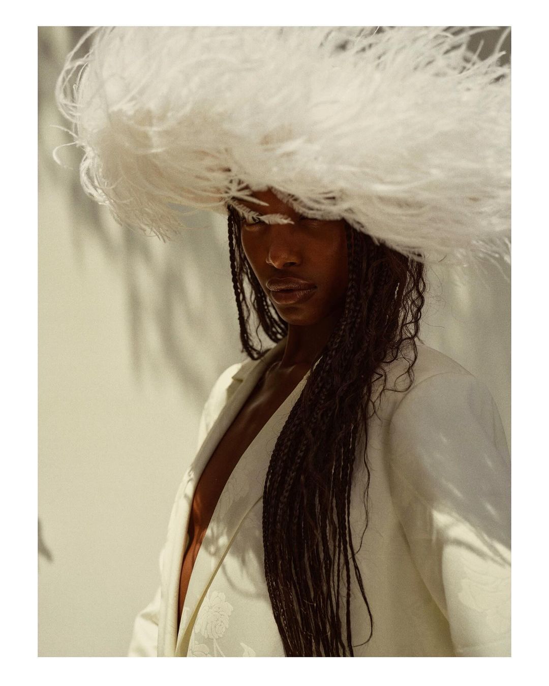 Woman in oversized feather hat and white jacket poses against a sun-drenched wall, showcasing fashion and beauty in a portrait.