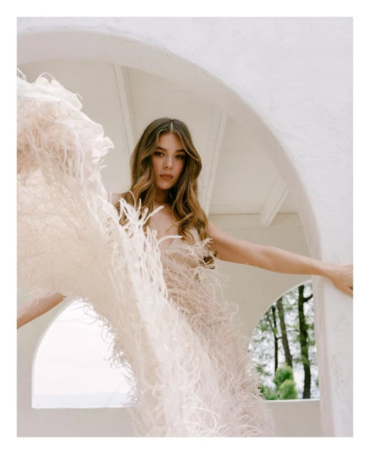 Young woman in feather dress posing near arched structure, medium shot. Fashion portrait against blurred green foliage backdrop.