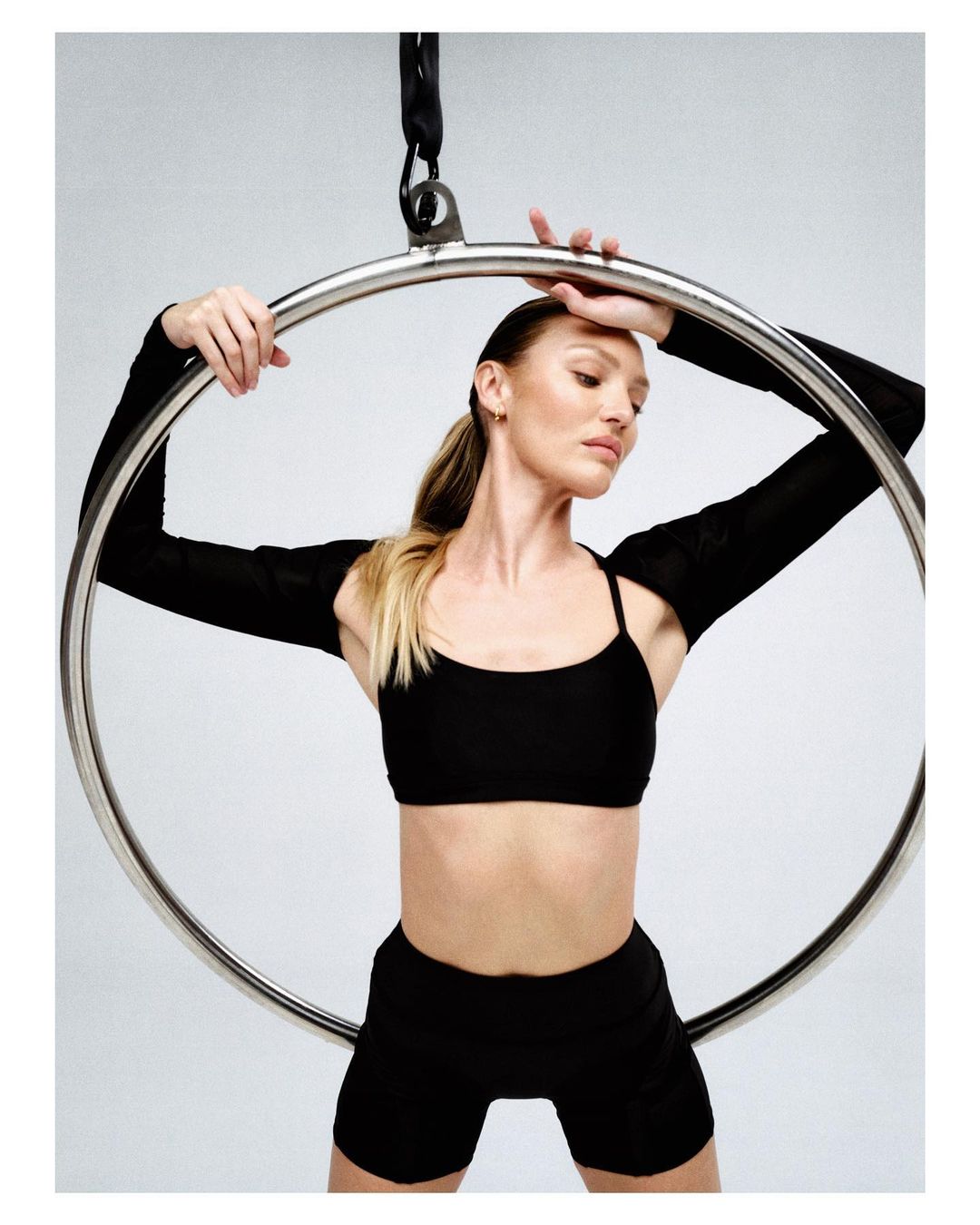 Adult woman in black workout clothes holding an aerial hoop with a neutral expression in a photo studio with a grey background.