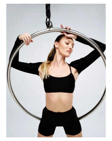 Adult woman in black workout clothes holding an aerial hoop with a neutral expression in a photo studio with a grey background.
