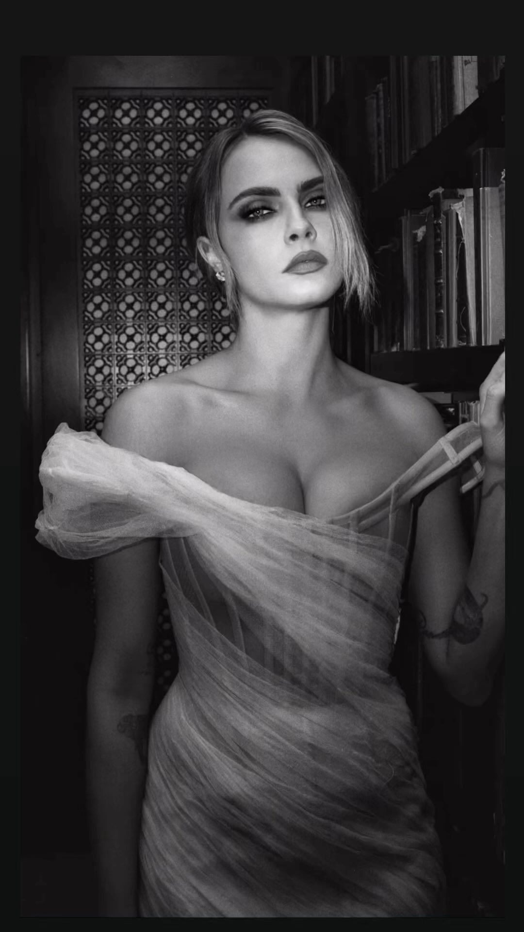 Portrait of Cara Delevingne in an off-the-shoulder tulle dress inside a library with bookshelves.