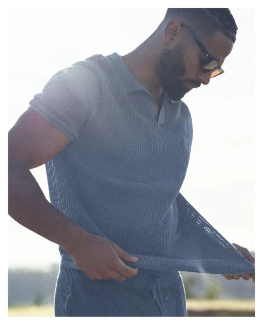 Adult male wearing sunglasses and a blue knit short-sleeve shirt with a collar, partially pulling on the shirt, outdoors in bright light.