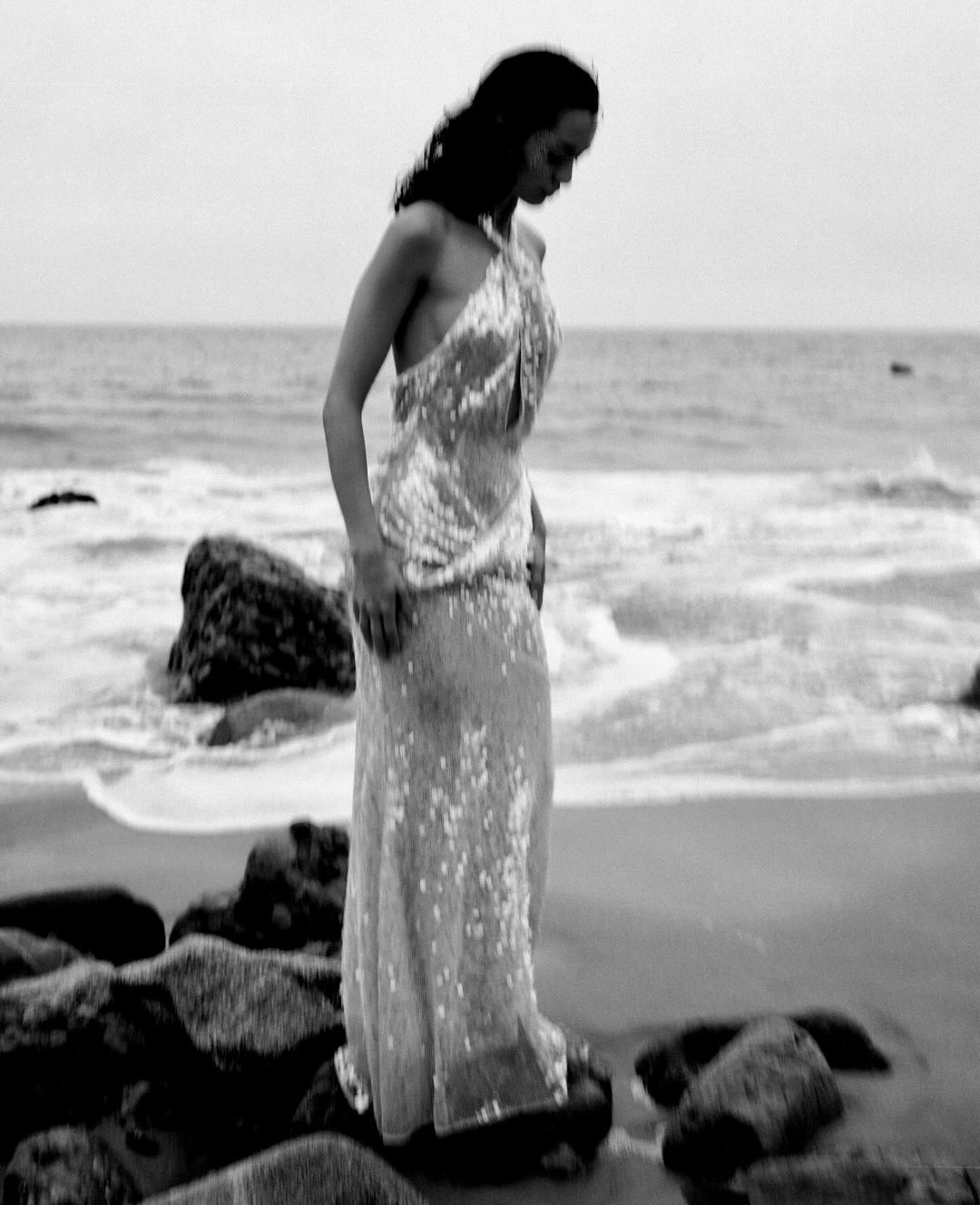 A woman wearing a shimmering halter gown standing on rocky shoreline by the ocean