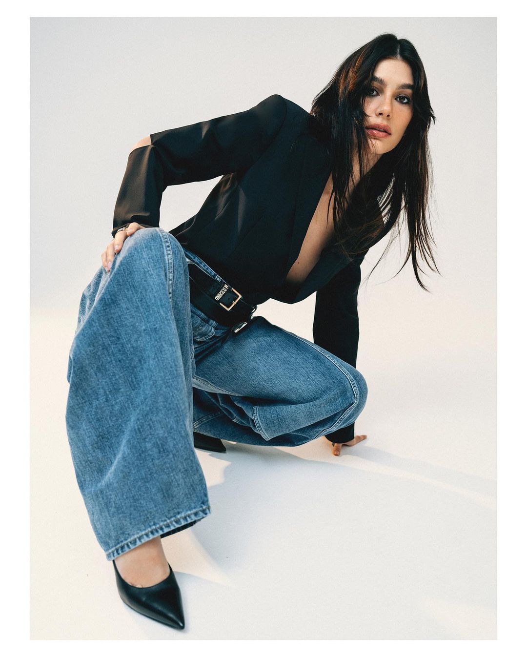 Brunette woman in a black blazer, loose jeans, and heels poses crouching in a studio with a neutral background, fashion editorial style.