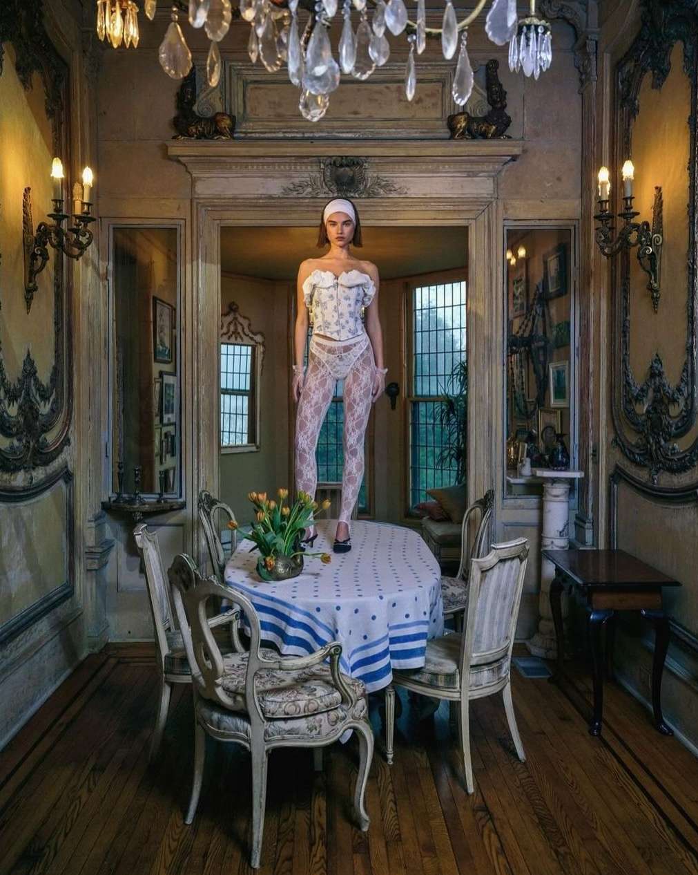 Woman in lingerie standing on a table with yellow tulips in an ornately decorated room with a chandelier and a doorway in the background.