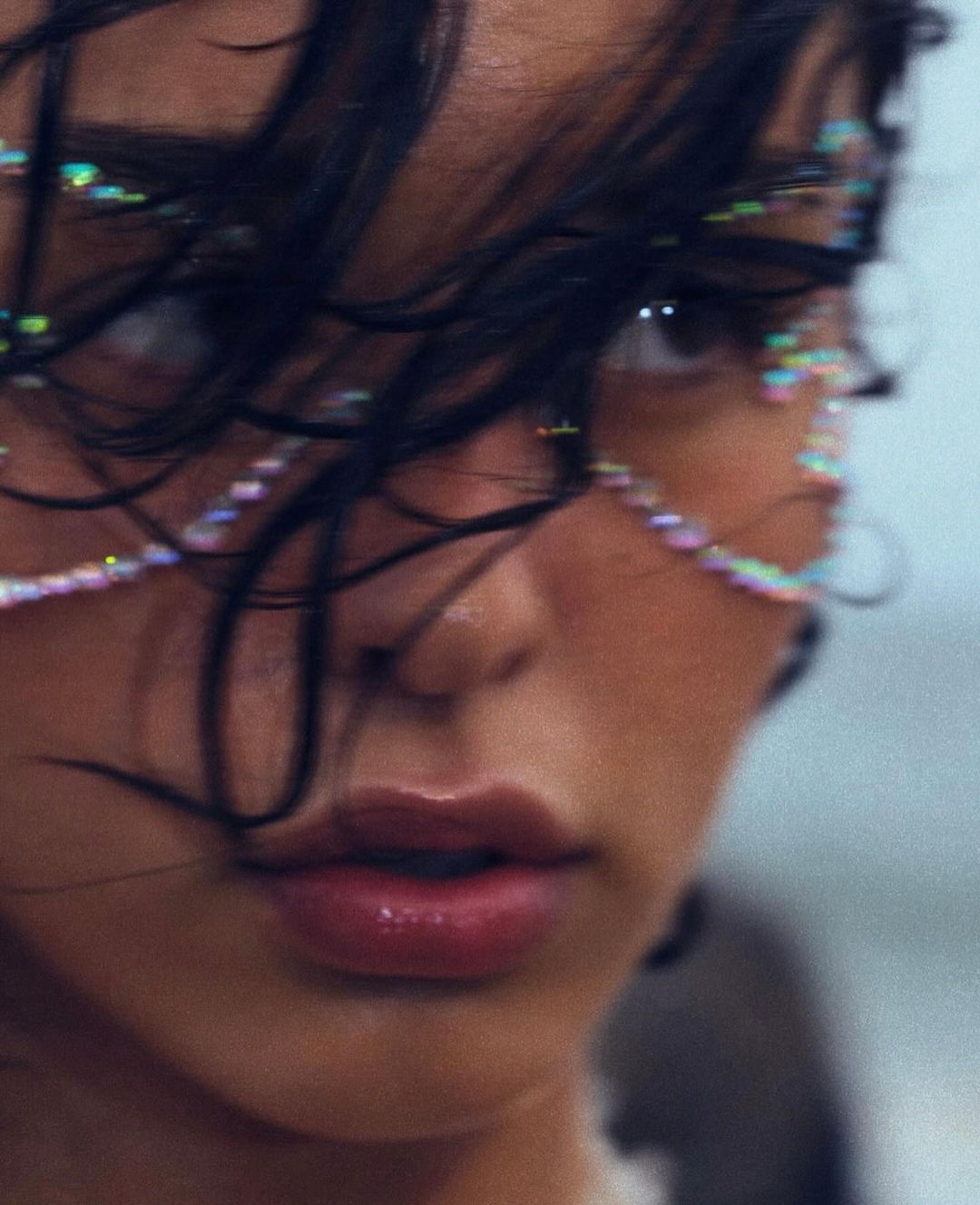 Close-up shot of a woman with dark hair and iridescent beaded strands across her face, standing outdoors with a blurred background.