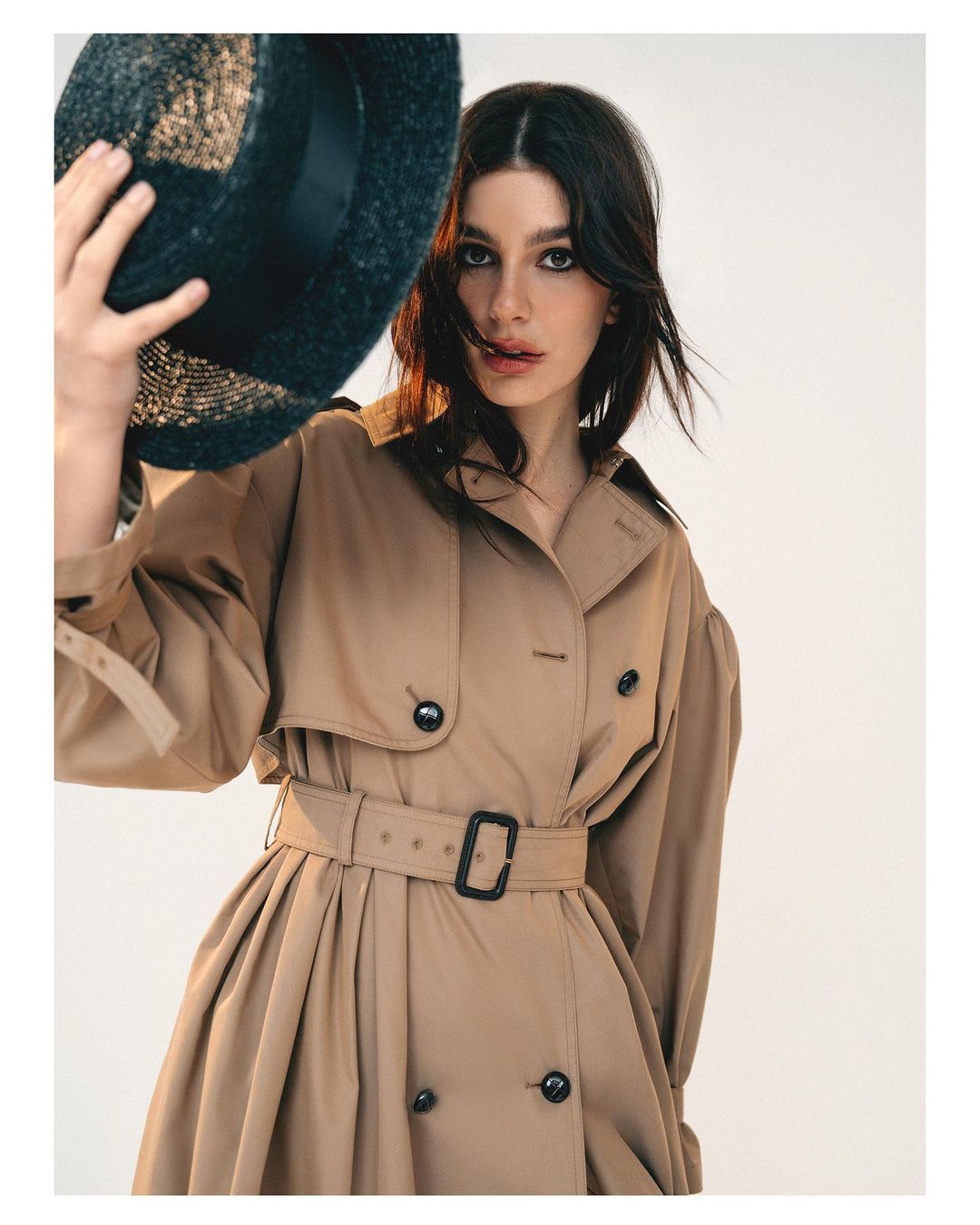 Woman wearing beige trench coat and holding dark straw hat by photographer Bryan Rodner Carr against a white backdrop