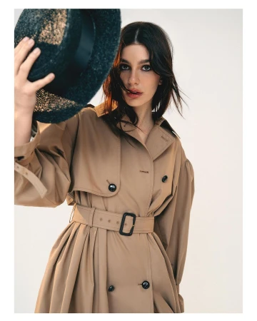 Woman wearing beige trench coat and holding dark straw hat by photographer Bryan Rodner Carr against a white backdrop