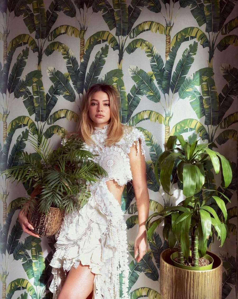 Indoor portrait of a blonde woman in a white crochet dress, holding a potted plant, against a backdrop of a leafy wallpaper.
