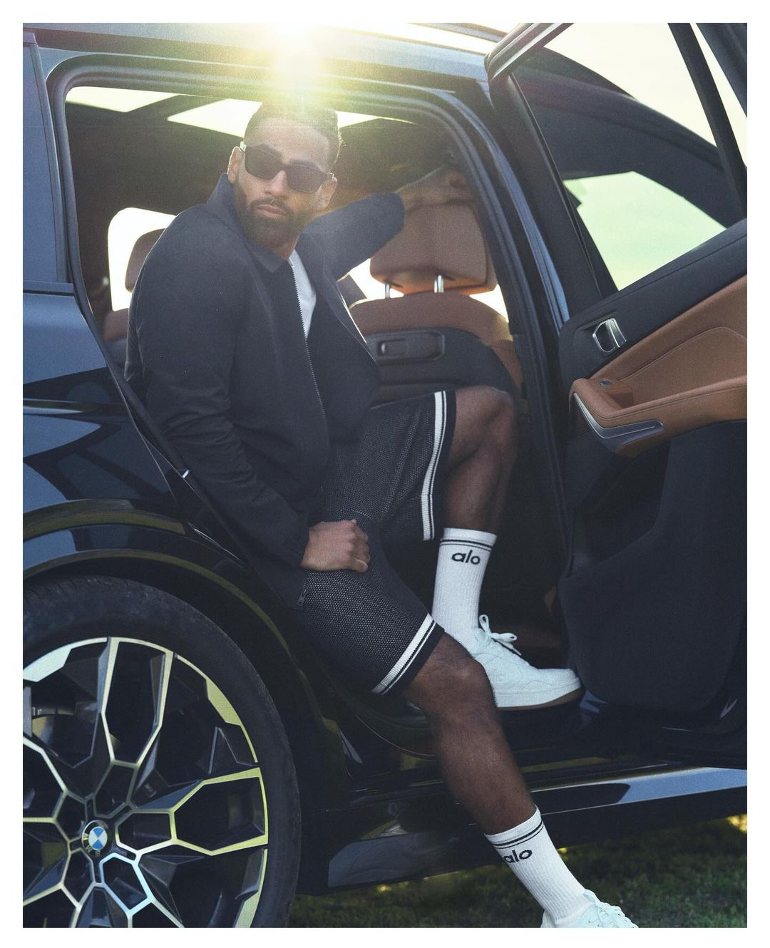 Athletic man exits a black SUV, wearing a black jacket, shorts with white stripes, white socks with a logo, and white sneakers.