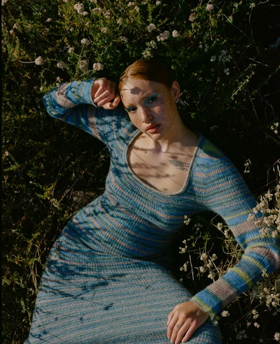Woman with red hair and blue dress lying in field of small white flowers in an outdoor setting, viewed from an eye-level angle.