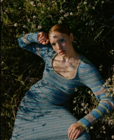 Woman with red hair and blue dress lying in field of small white flowers in an outdoor setting, viewed from an eye-level angle.