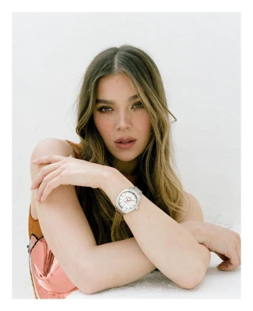 Portrait of a woman with long blonde hair wearing a silver wristwatch, shot in a bright photo studio with a light background.