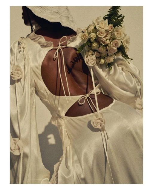 A woman with dark skin wearing a white dress and hat is seen from the back, holding white roses in a modern fashion shot.
