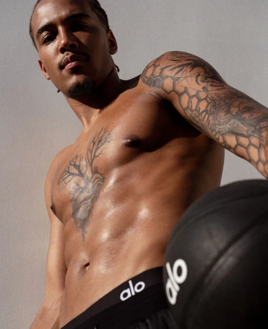 Shirtless man with tattoos poses with a black ball in a close-up shot against a neutral background, wearing Alo branded bottoms.