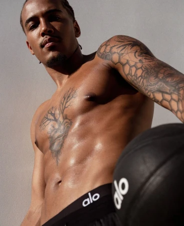Shirtless man with tattoos poses with a black ball in a close-up shot against a neutral background, wearing Alo branded bottoms.